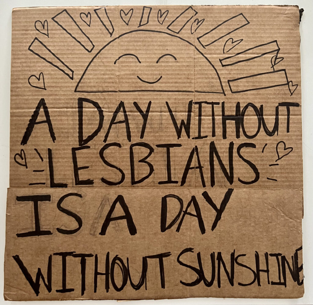 Cardboard sign brought to 2022 pride parade, this side "A Day Without Lesbians is a Day Without Sunshine" recreated from the San Francisco Gay Freedom Parade 1979 in black sharpie.