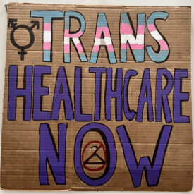 Cardboard sign brought to 2022 pride parade, this side "TRANS HEALTHCARE NOW" painted with acrylic paint with with anti abortion and trans symbols.