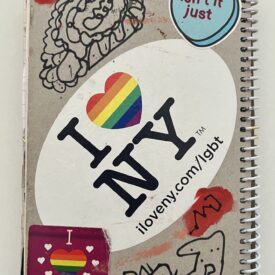 Spiral notebook back with a large "I Heart NY" sticker with a rainbow heart in the center. Also black abstract squiggles, "I Heart Inclusive Schools," and "Isn't it Just" inside a blue heart.