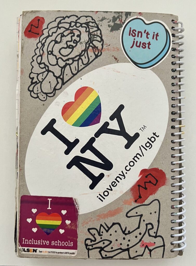 Spiral notebook back with a large "I Heart NY" sticker with a rainbow heart in the center. Also black abstract squiggles, "I Heart Inclusive Schools," and "Isn't it Just" inside a blue heart.
