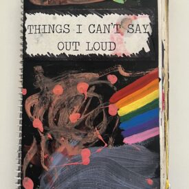 Spiral notebook with paint splatters, a painted rainbow, and "Things I Can't Say Aloud" written on a black background.