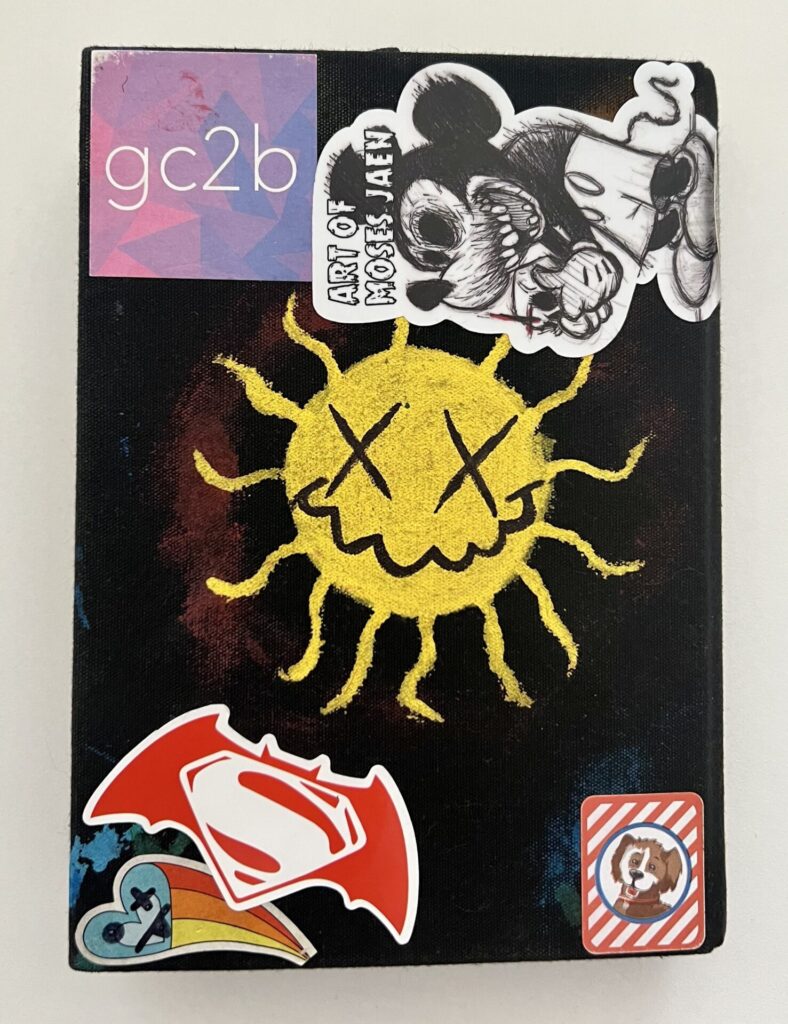 Black colored journal back with gc2b sticker, Mickey Mouse with "Art of Moses Jaen, drawing of a sun with X shaped eyes, Superman symbol inside Batman wings, a heart with trailing colors, and a cartoon dog surrounded by red and white stripes.